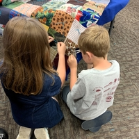 young boy and girl tie squares together kneeling on the ground