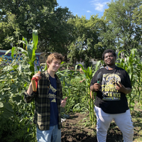 Two youth stand holding gardening tools surrounded by sunflowers