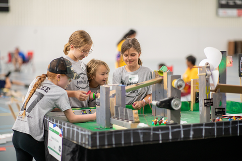 Four girls wearing matching gray team t-shirts work on their Rube Goldberg machine.