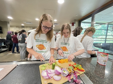 Two teen girls wearing 4-H YELLO t-shirts and cutting a variety of vegetables on wooden cutting board.