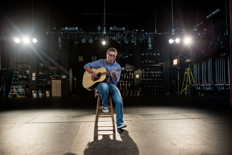 Boy on a stool playing a guitar.