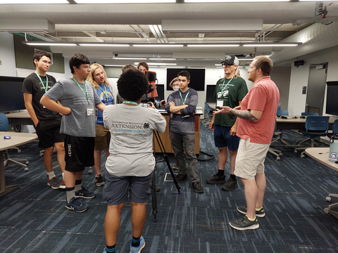 A group of youth standing around a video camera on a tripod as a man talks. 