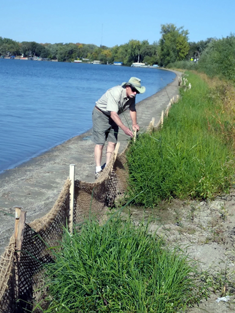 A man in a hat and sunglasses checks a net fence along a lakeshore. Sand is on the lakeside and vegetation is on the farther side of the fence.