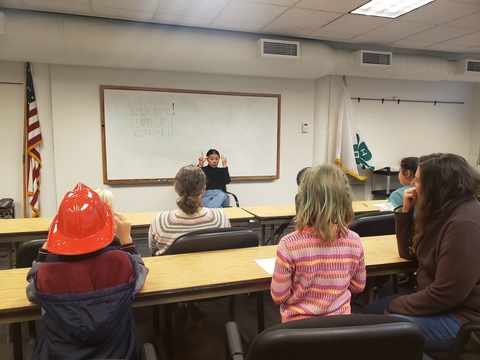 A woman doing sign language in front of a classroom of kids and adults.