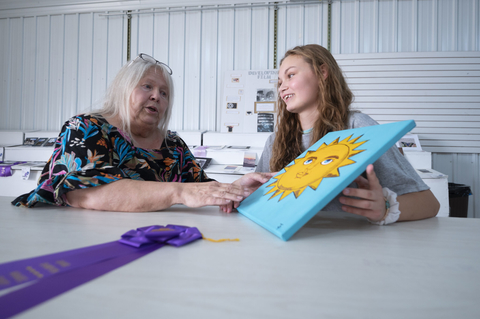 An older woman talking to a young girl about a painting of a sun she is holding.