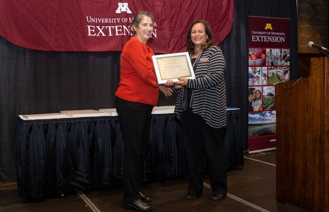 Dean Bev Durgan hands award plaque to Carol Cardona on a stage. They are smiling facing the audience.