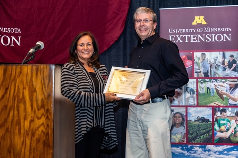 Dean Bev Durgan and Brian hold award plaque between them on a stage. Brian is wearing khakis and a black button-down shirt.