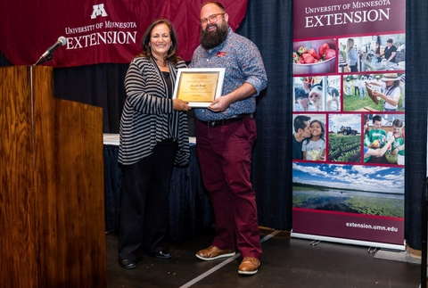 Dean Bev Durgan and Joe Rand hold award plaque between them on a stage. Joe has a huge dark beard and deep red pants.