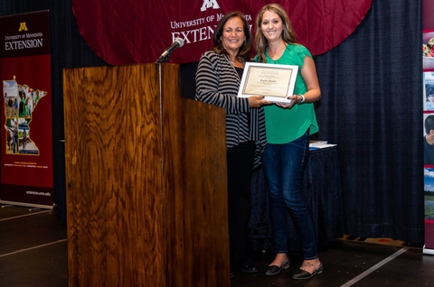 Dean Bev Durgan and Kayla hold award plaque between them on a stage. Kayla has on a green top and jeans.