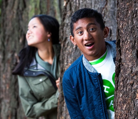 A boy and girl peeking out from behind trees.