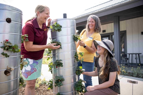 Three women look at each other and at the strawberries growing out of holes in an aluminum tower that is about their height.