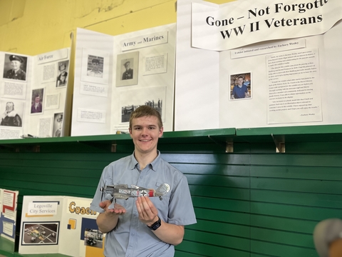 Zach W. holding his plane exhibit in front of a display board titled, "Gone - Not Forgotten - WWII Veterans."