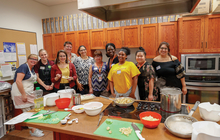 Group of people stand around kitchen counter with cooking materials.