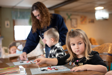 Parent helping two kids with art projects, baby in the background.