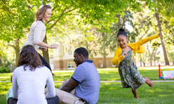 A woman playing outside with a girl while a man and a woman sit on the grass smiling.