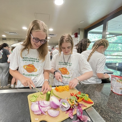 Two teen girls wearing 4-H YELLO t-shirts and cutting a variety of vegetables on wooden cutting board.