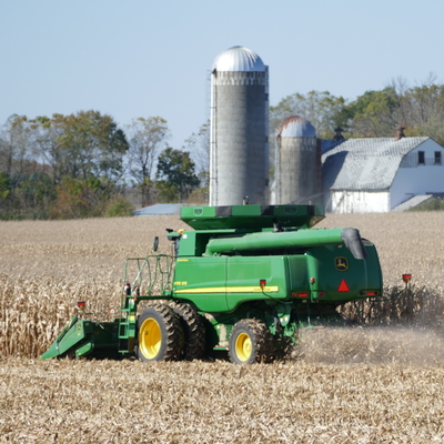 Harvester harvesting corn with a silo and farm in the background.