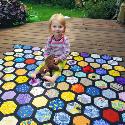 A girl sits on a quilt with a rainbow honeycomb design.